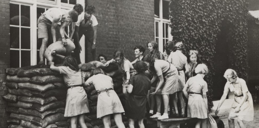 Black and white image of Girl Guides build a sandbag barrier against a building
